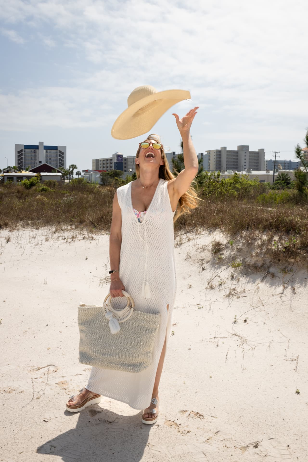 Model tossing a straw hat in a white knit maxi dress on the dunes