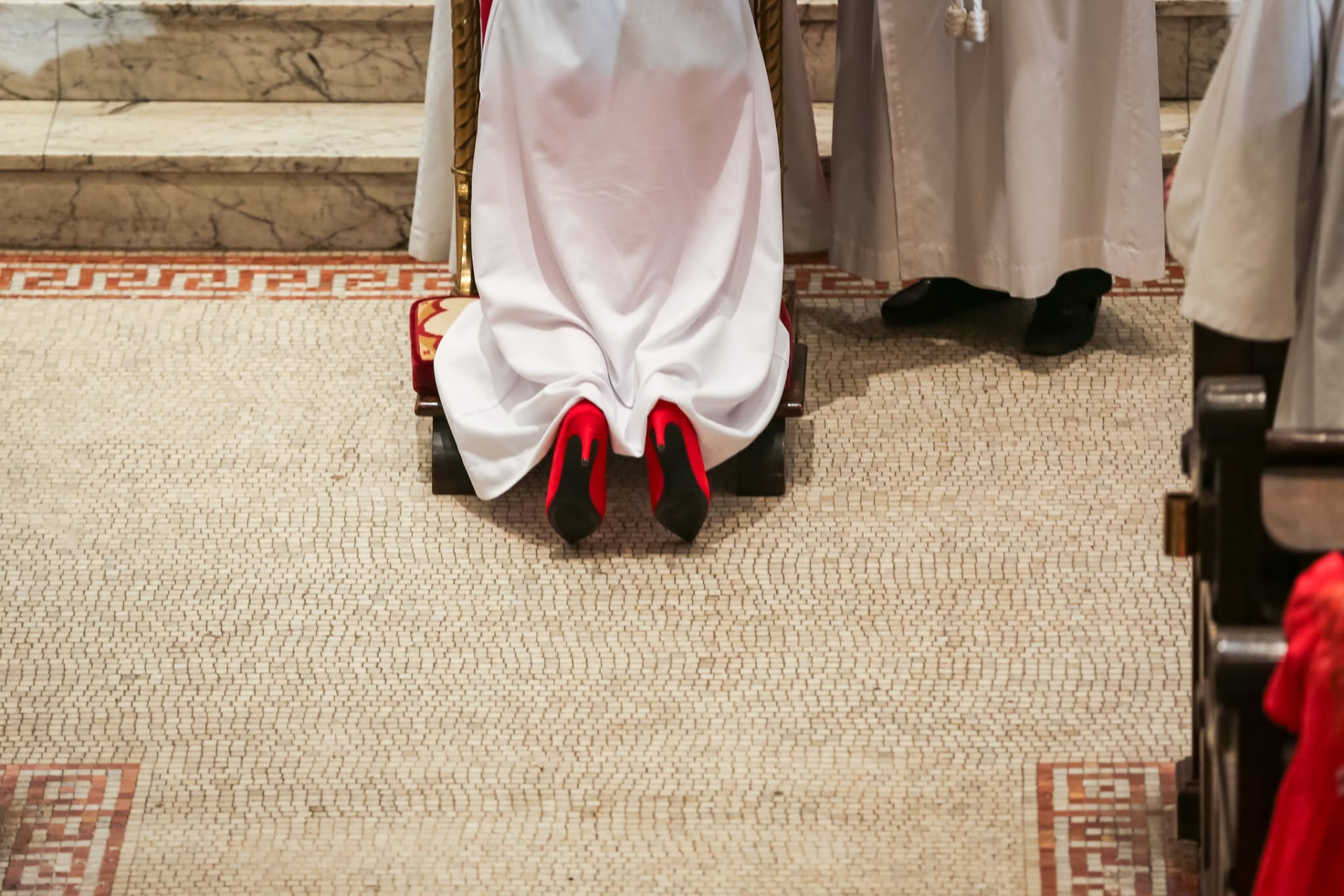 A kneeling figure in white vestments wearing red high-heeled pumps on a church kneeler