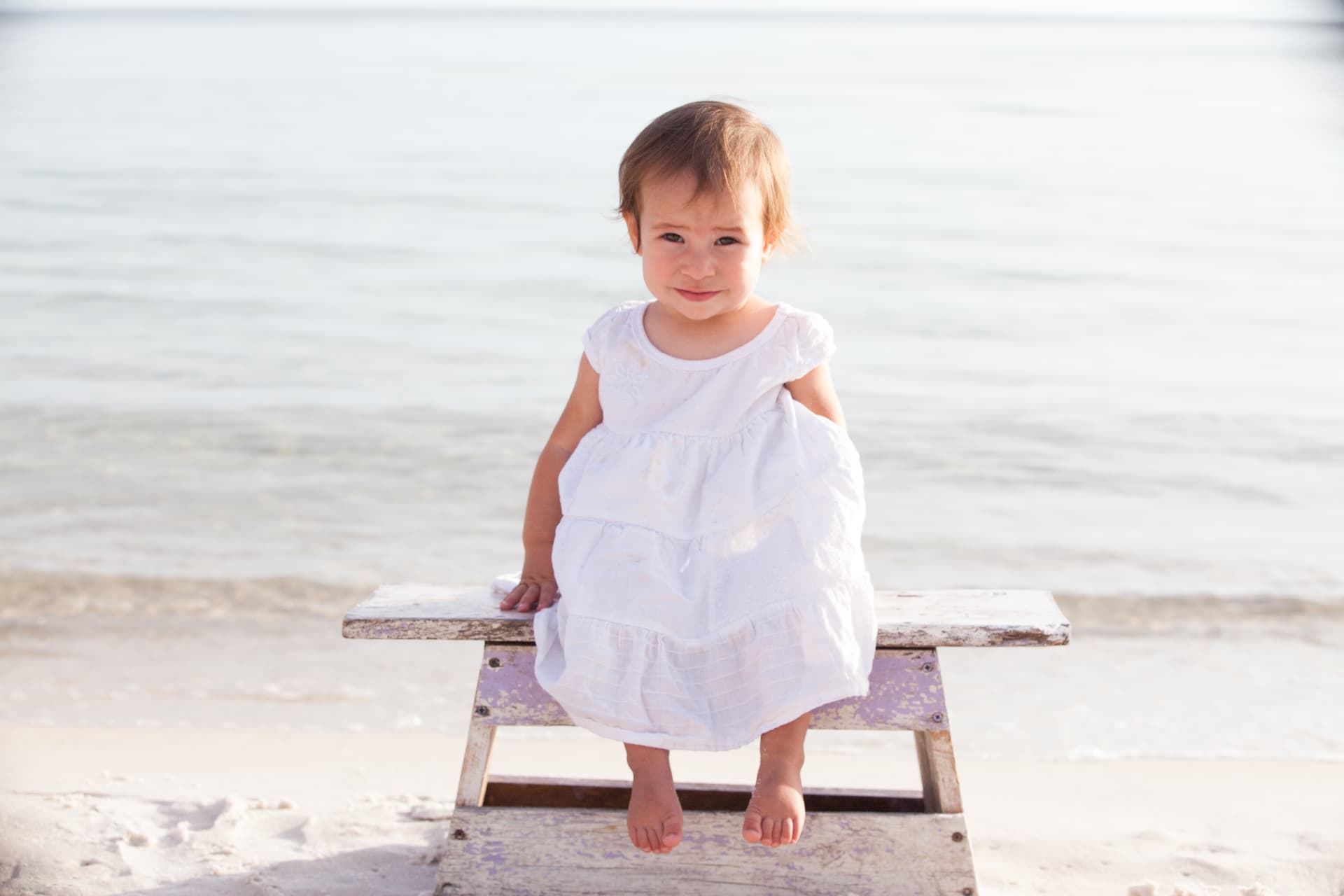 Toddler in a white dress on a bench at the water's edge