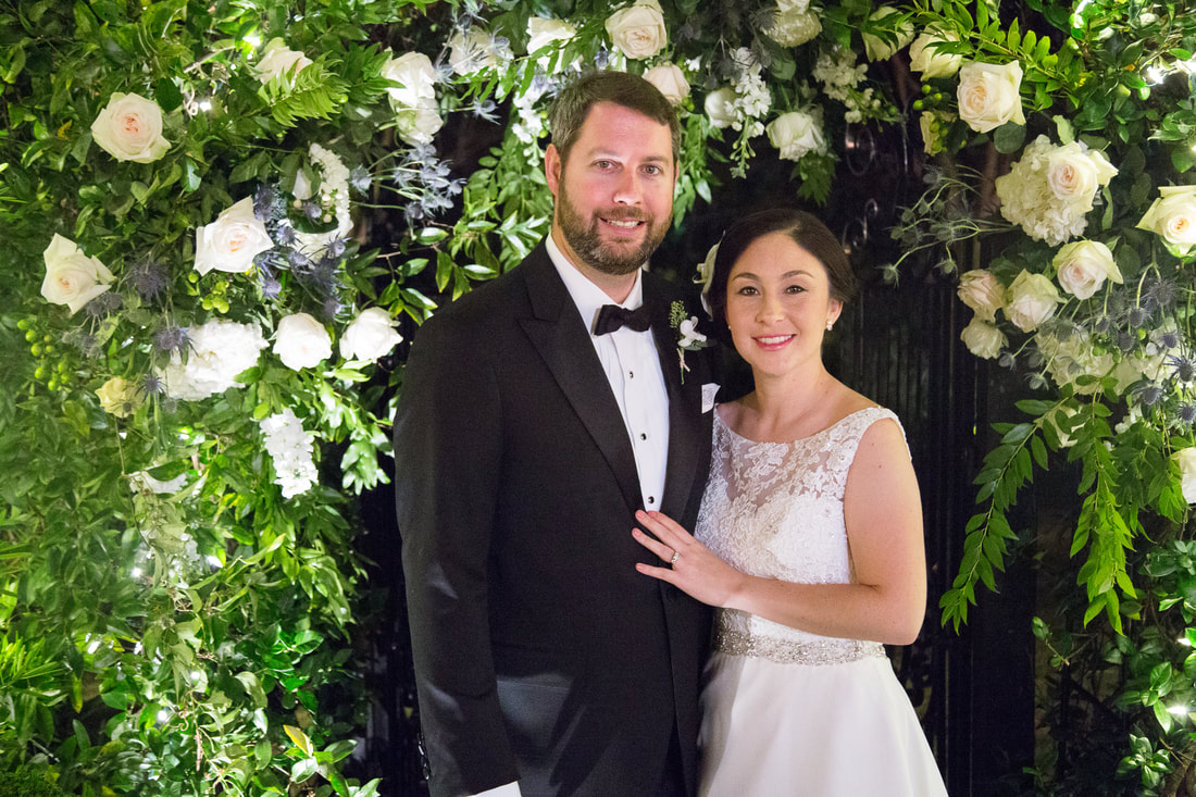 Bride and groom beneath a white rose and greenery arch