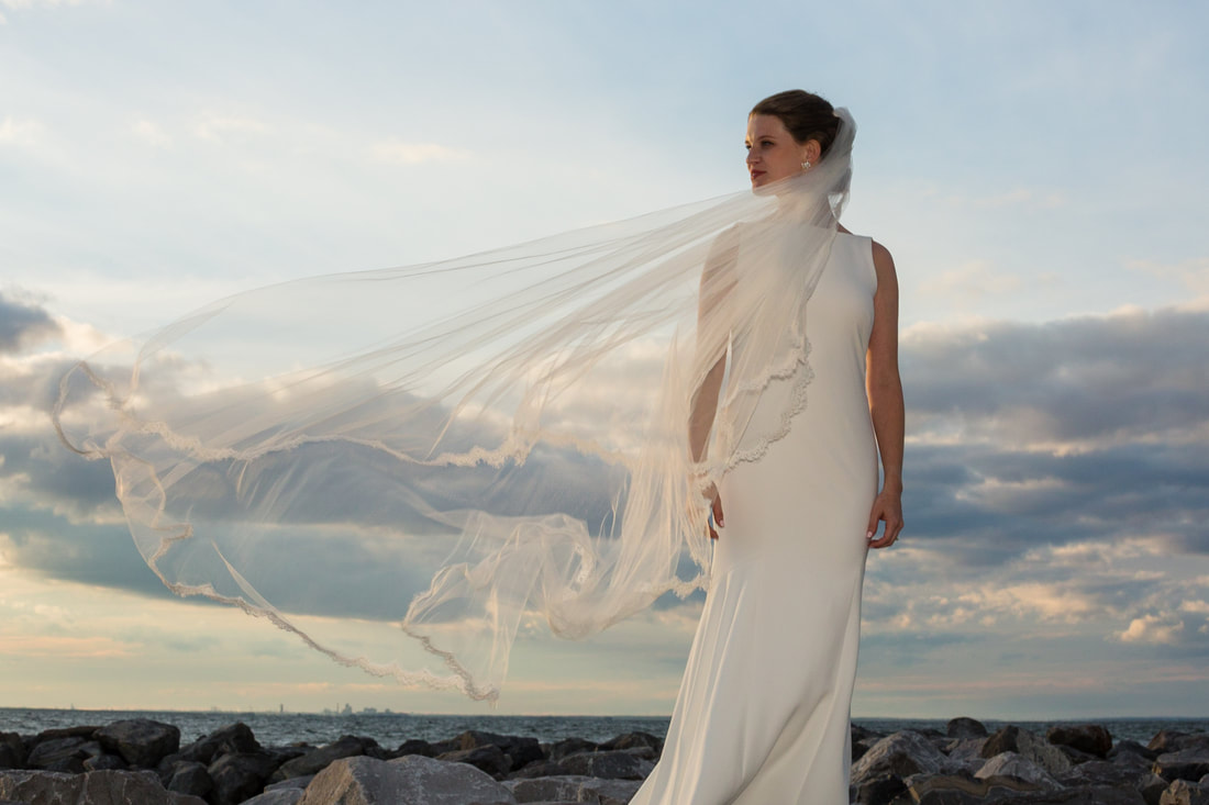 Bride with her veil catching the wind by the water at sunset